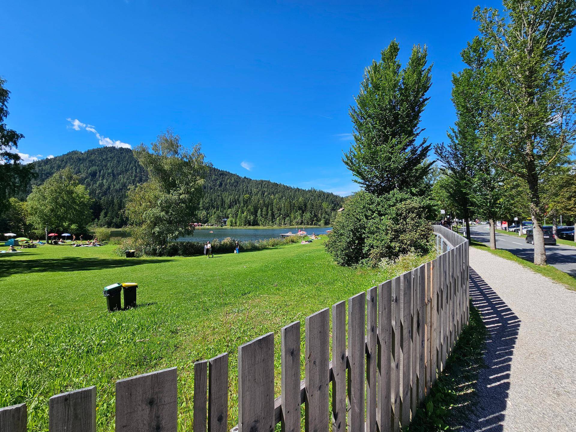 Blick auf einen See und bewaldete Hügel von einer grünen Wiese mit einem Holzzaun im Vordergrund.