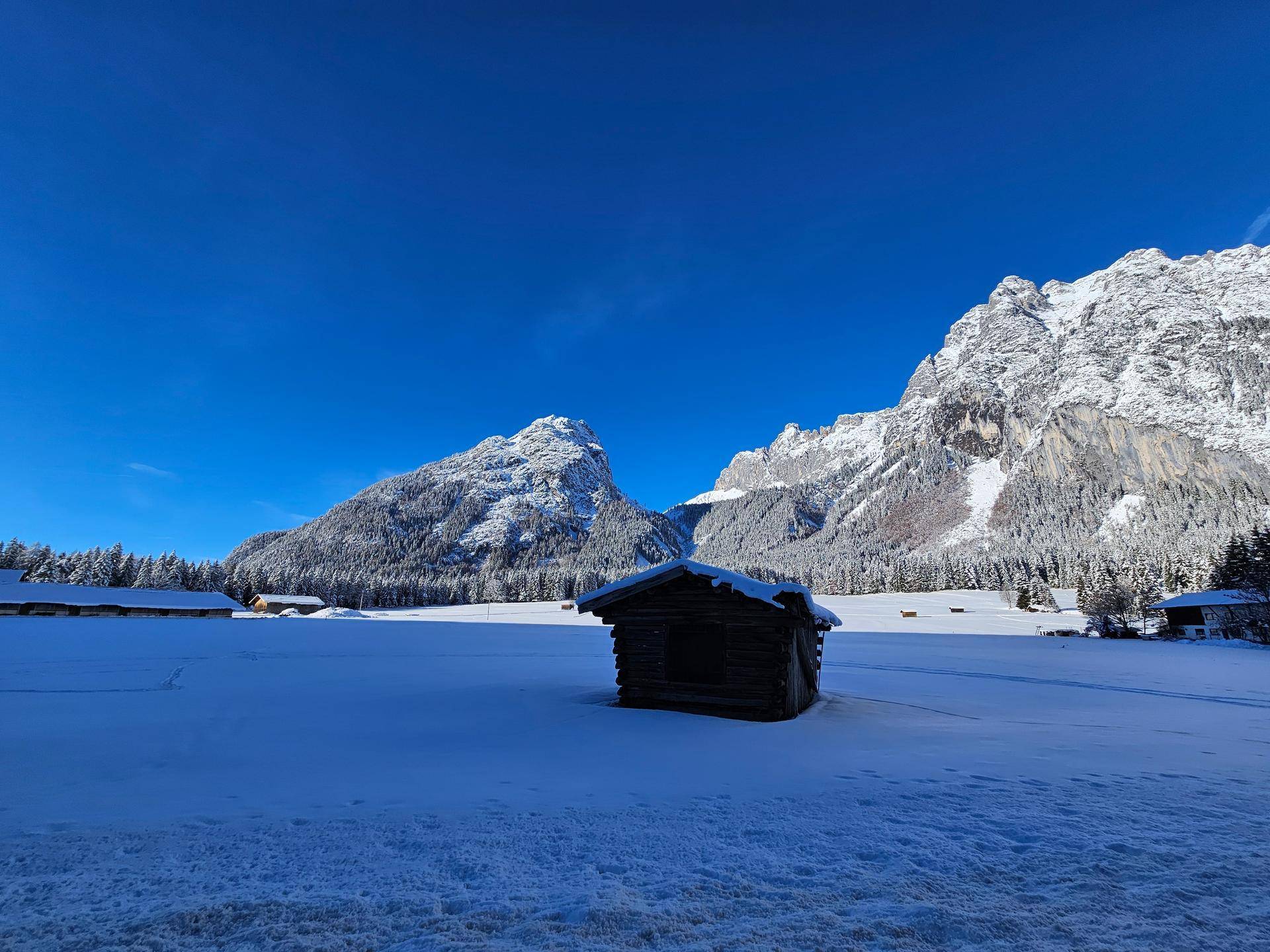 Eine kleine Holzhütte steht in einer weiten, verschneiten Ebene vor imposanten, schneebedeckten Bergen unter blauem Himmel.