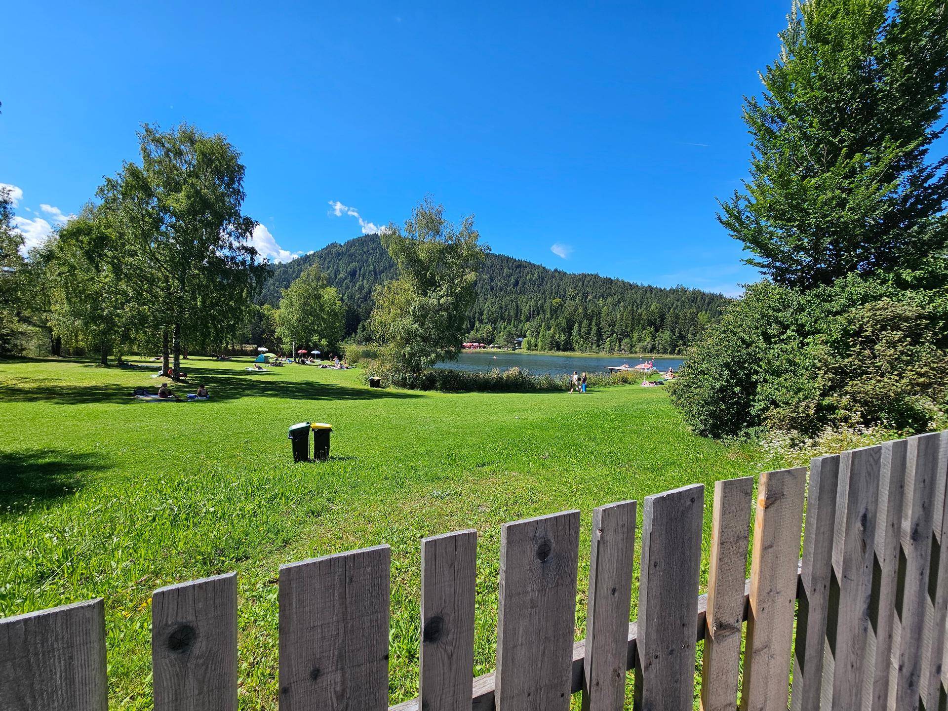Panoramablick auf einen ruhigen See, umgeben von grünen Wiesen und Wäldern unter blauem Himmel.