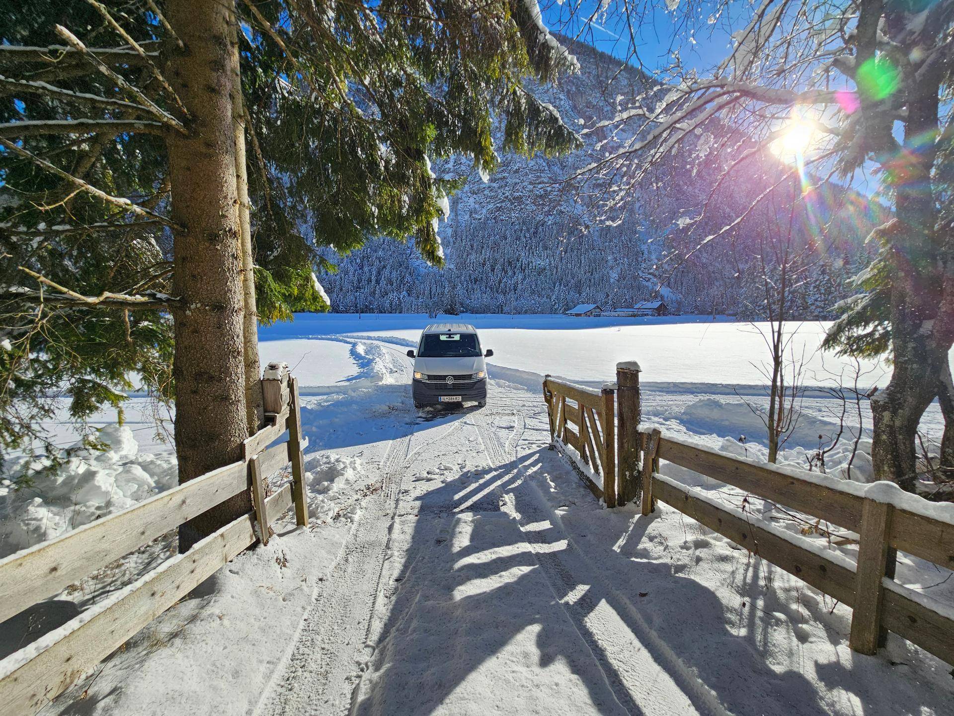 Ein verschneiter Weg führt durch eine Winterlandschaft zu einem weißen Van, umgeben von Bäumen und Bergen.