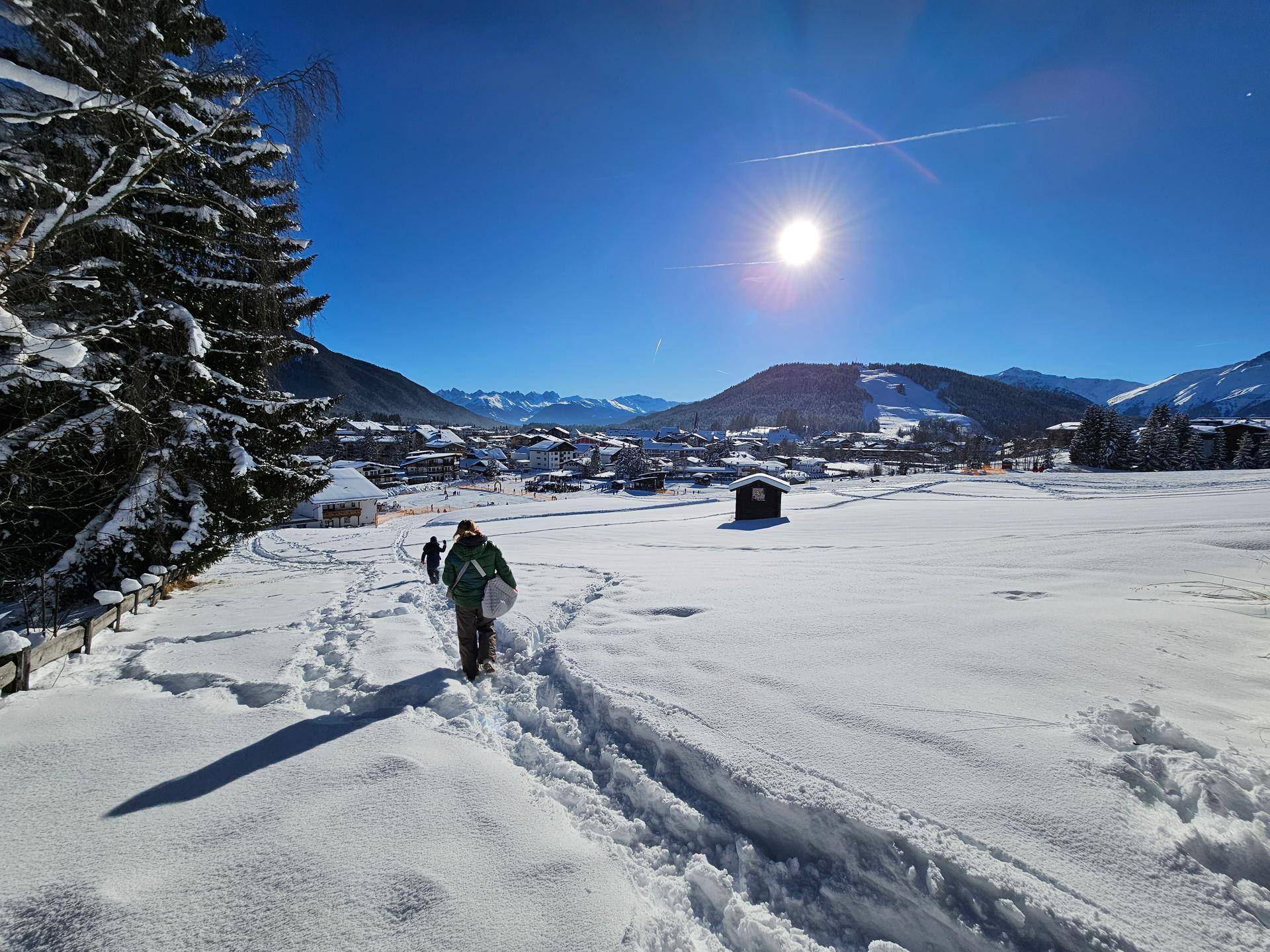 Person stapft durch tiefen Schnee auf einem Winterweg mit einem Dorf und Bergen im Hintergrund.