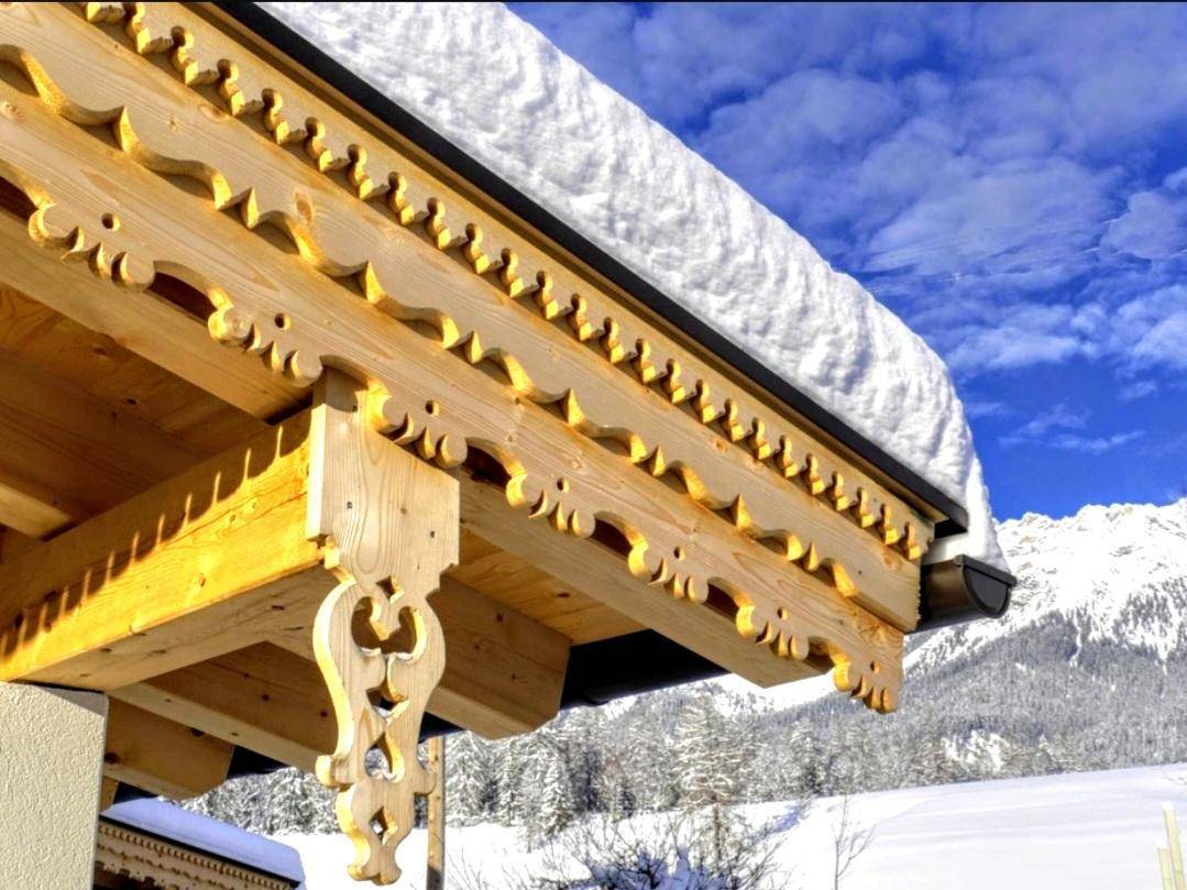 Detailaufnahme von kunstvoll geschnitzten Holzbalken unter einem verschneiten Dach mit Bergblick.