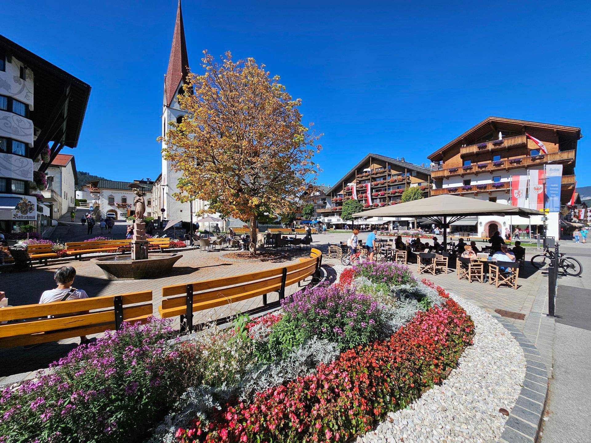 Belebter Stadtplatz mit einer Kirche, einem Brunnen, bunten Blumenbeeten und Außengastronomie.
