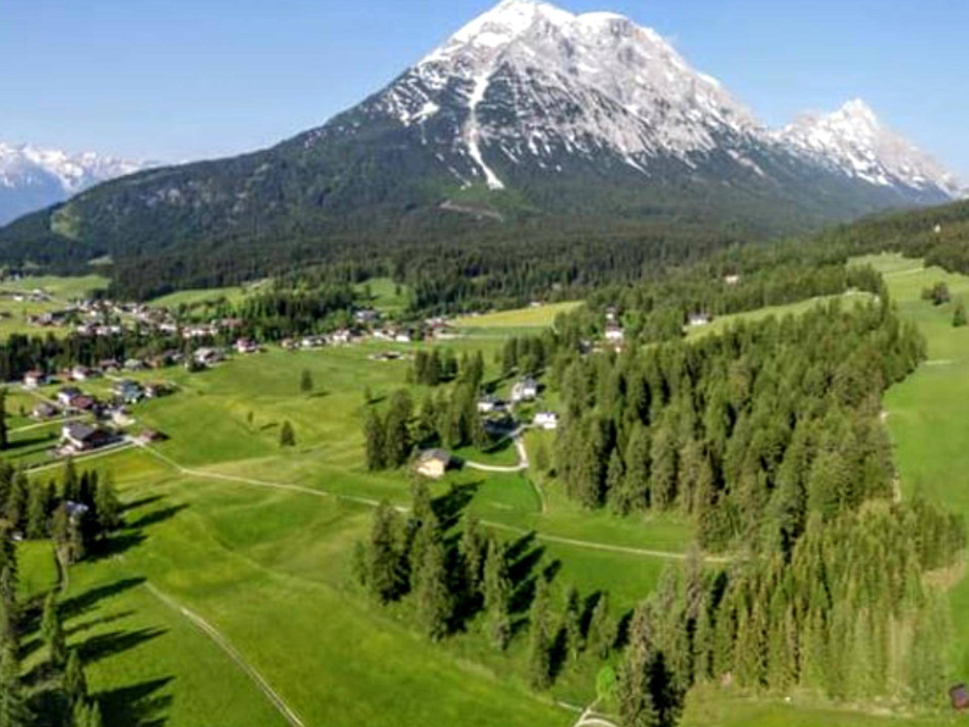 Luftaufnahme eines Dorfes umgeben von Wäldern und grünen Wiesen mit einem majestätischen Berg im Hintergrund.