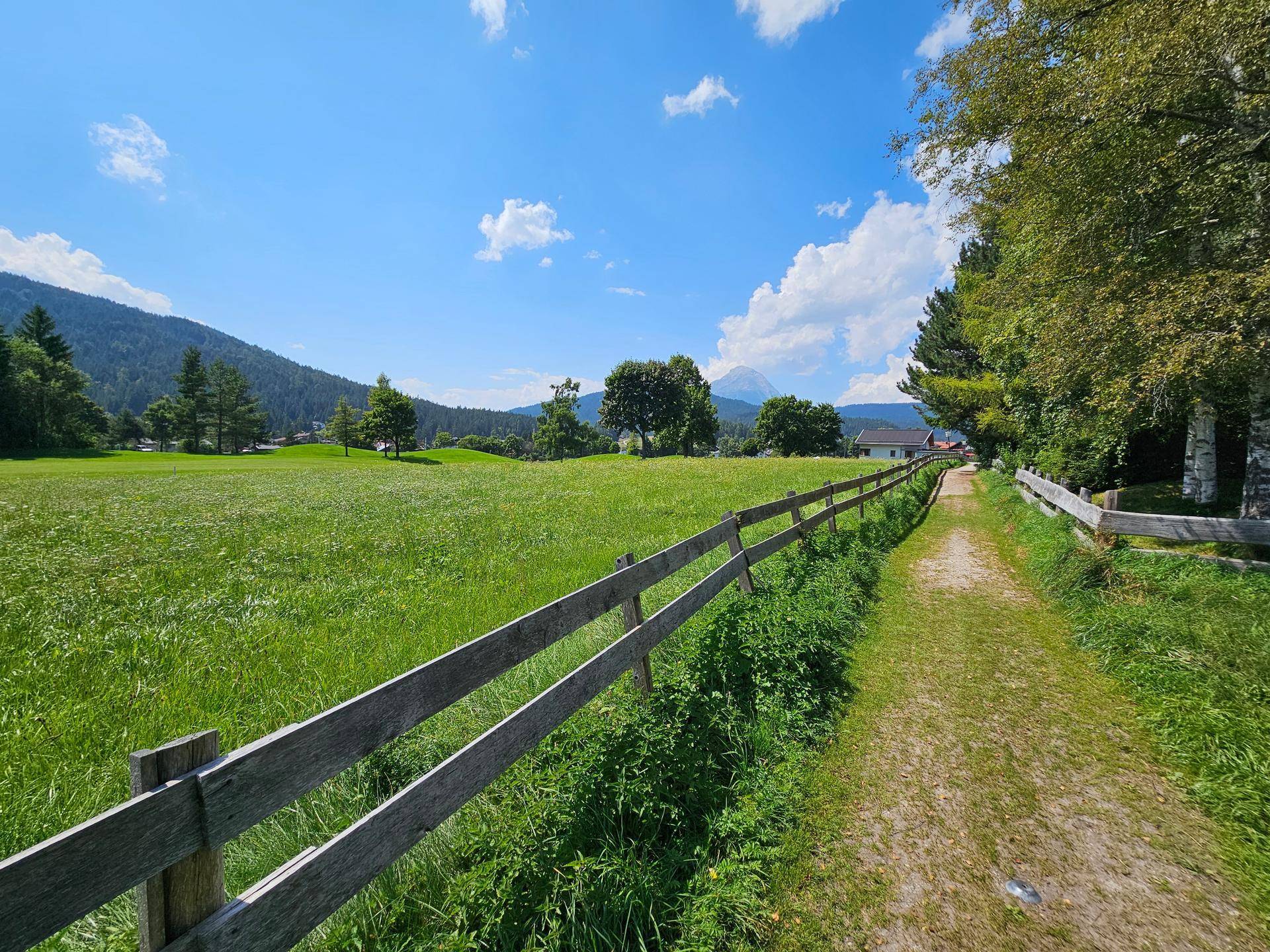 Feldweg entlang eines Holzzauns mit grünen Wiesen, Bäumen und Bergen in der Ferne.