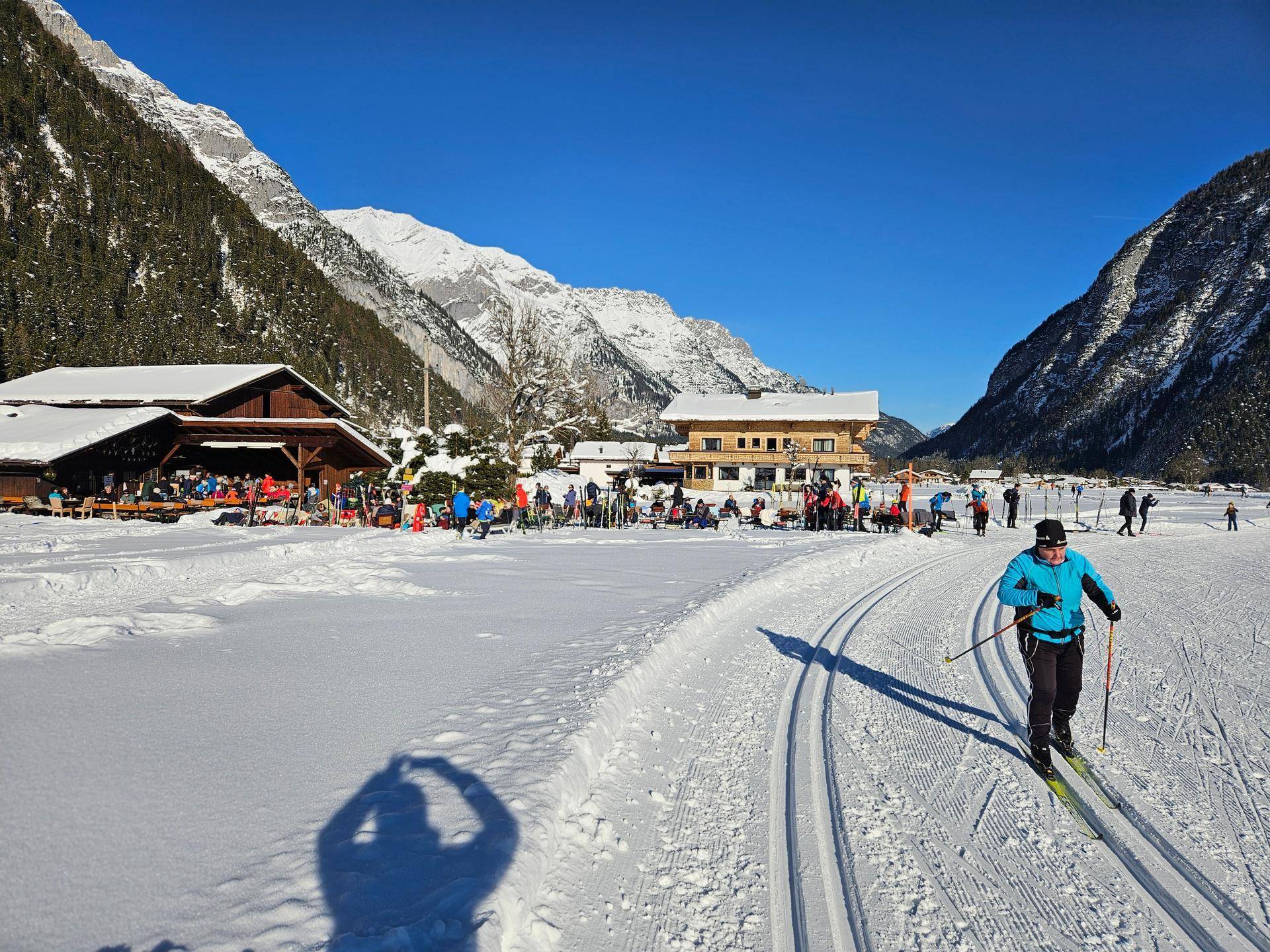 Zahlreiche Langläufer auf Loipen in einem verschneiten Tal mit Gebäuden und Bergen.
