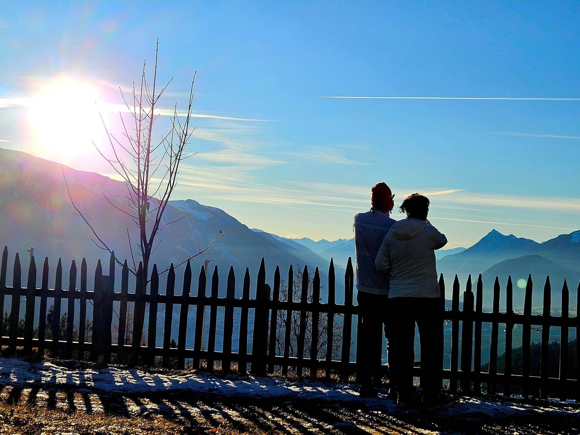 Zwei Personen betrachten einen weiten Bergblick über einen Holzzaun bei Sonnenaufgang oder Sonnenuntergang.