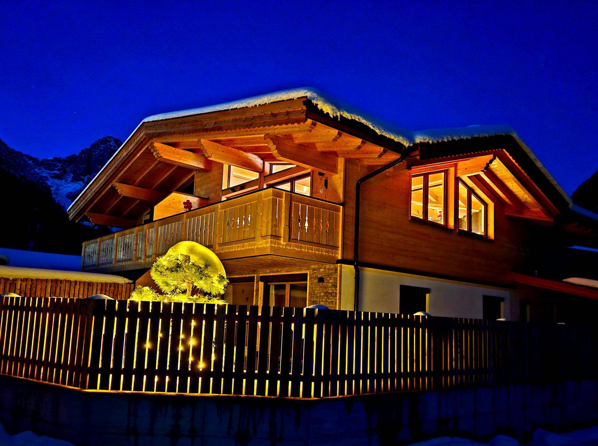 Gemütliches Holzhaus im Winter bei Nacht, mit beleuchtetem Balkon und verschneitem Dach.