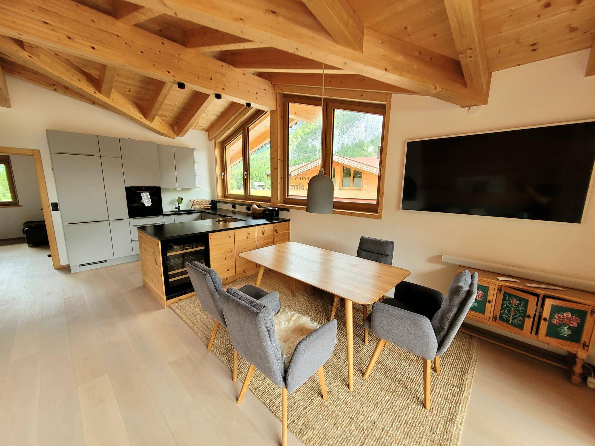 Helle offene Küche mit Essbereich, grauen Schränken und einem Holztisch unter Holzbalkendecke.