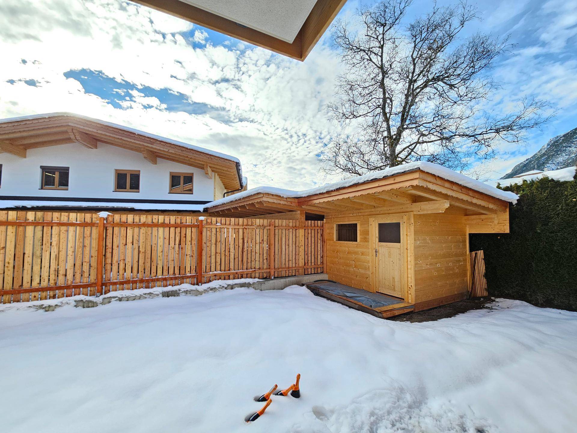 Holzgartenhaus in einem verschneiten Garten, umgeben von einem Holzzaun und einem Wohnhaus.