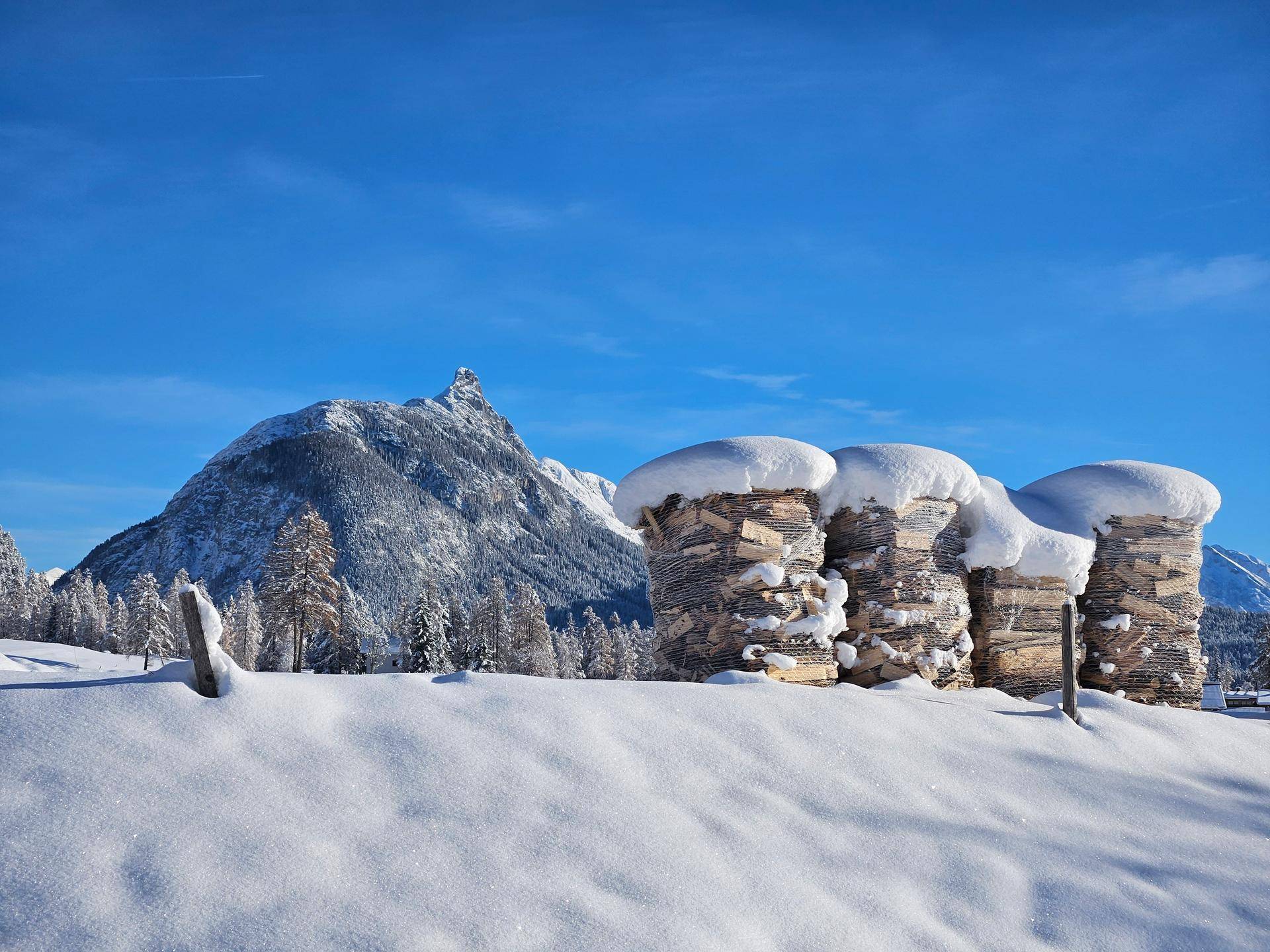 Drei schneebedeckte Holzstapel liegen vor einem imposanten, schneebedeckten Berg unter strahlend blauem Himmel.