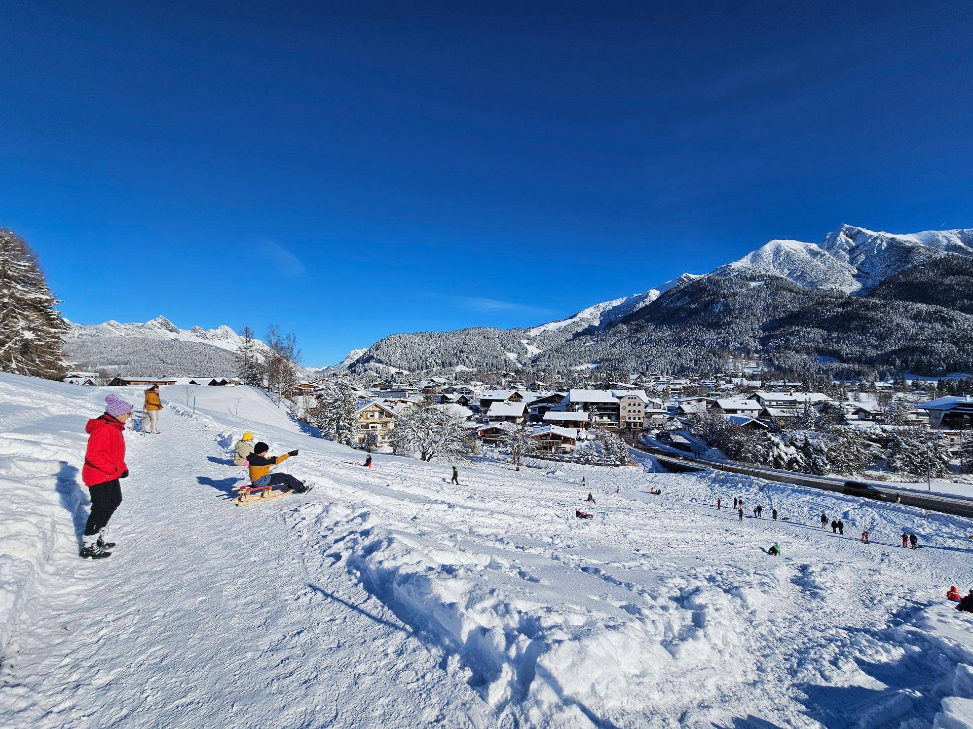 Kinder und Erwachsene rodeln einen verschneiten Hügel hinunter mit einem Dorf und Bergen im Hintergrund.