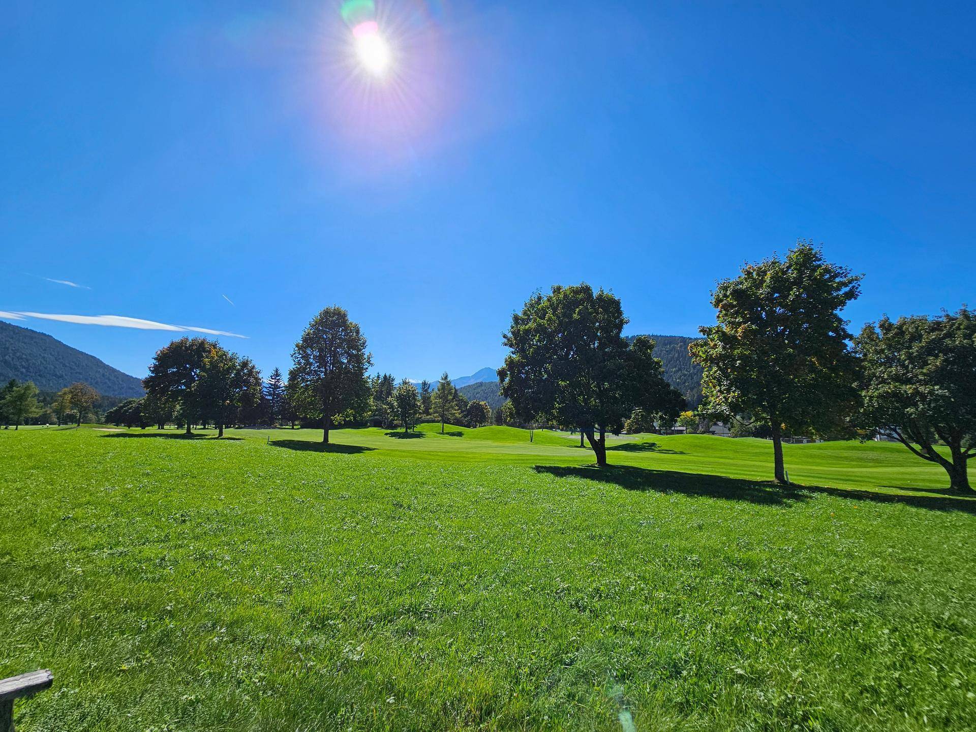 Weitläufiger grüner Golfplatz mit vereinzelten Bäumen und majestätischen Bergen im Hintergrund.