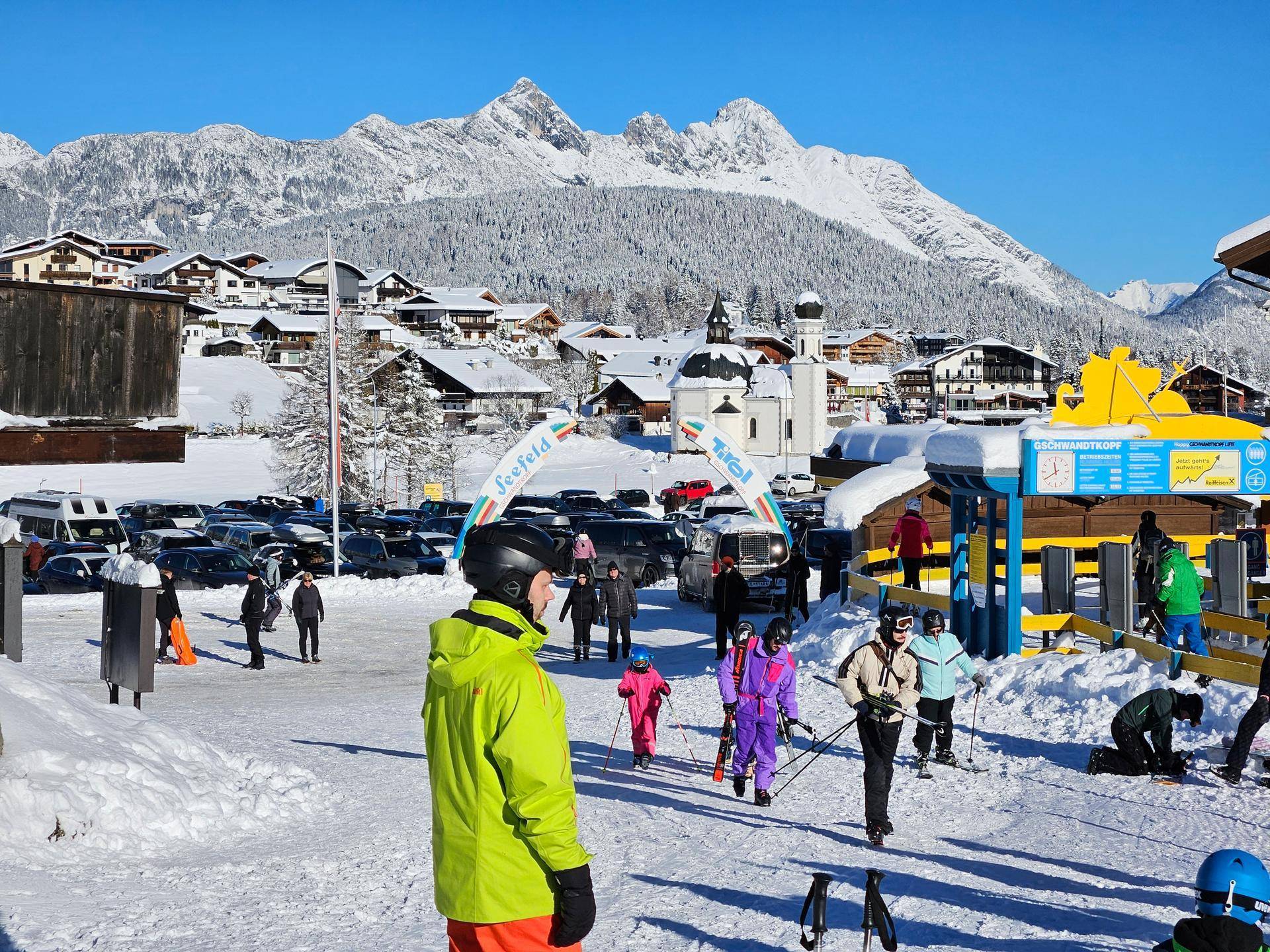 Belebter Skibereich in einem verschneiten Dorf mit Bergen im Hintergrund und vielen Skifahrern.