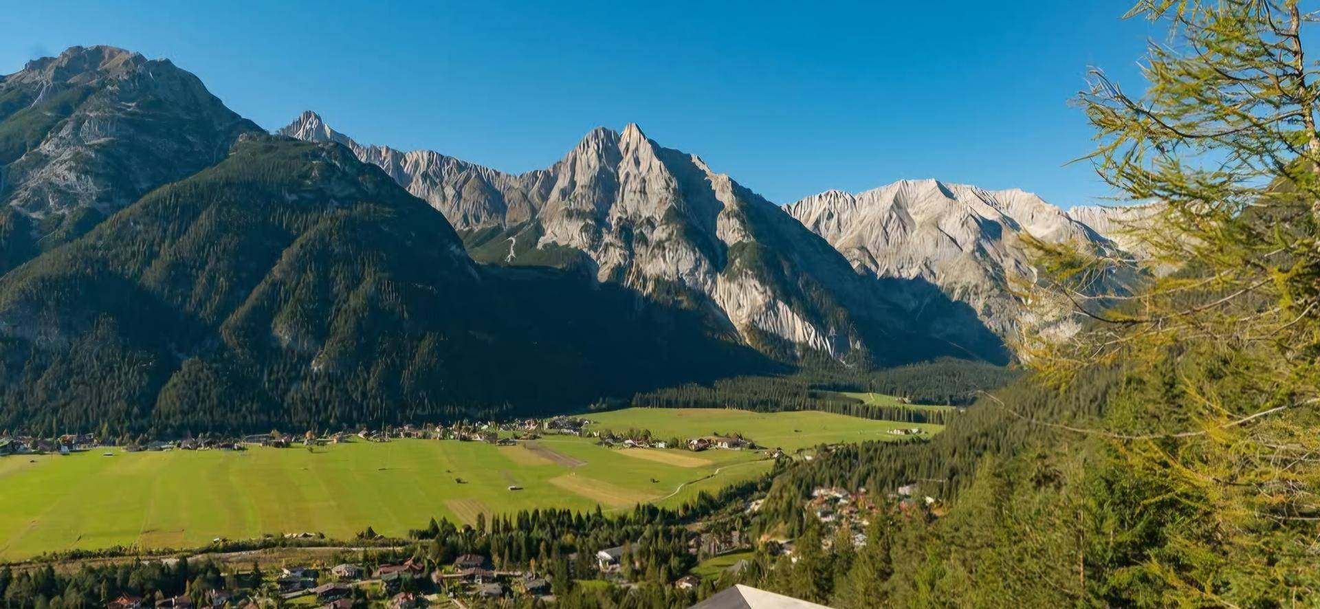 Weitläufiger Panoramablick über ein grünes Tal mit Dorf und majestätischen Bergen im Hintergrund.