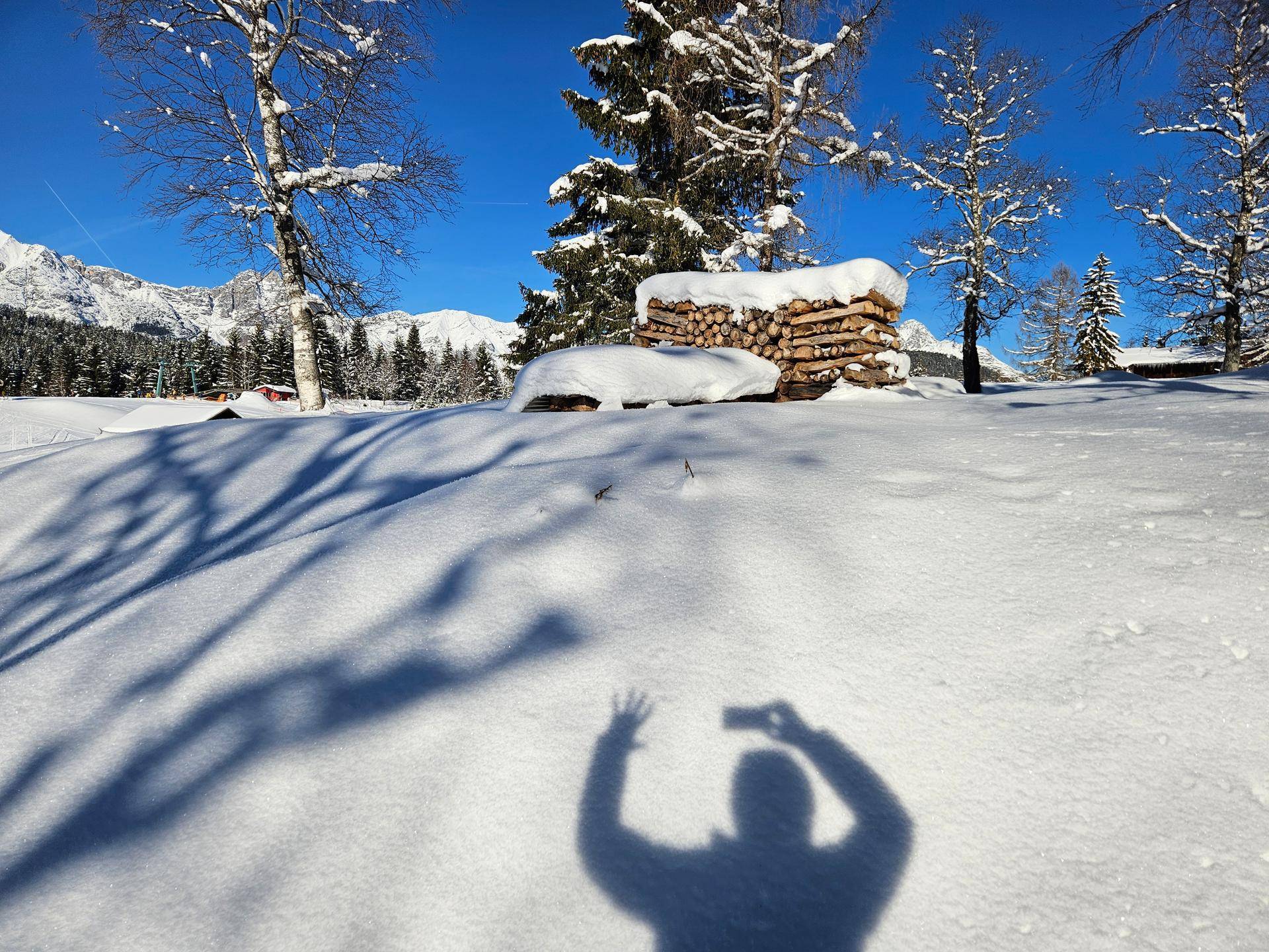 Verschneite Winterlandschaft mit einem Holzstapel und langen Schatten unter blauem Himmel.