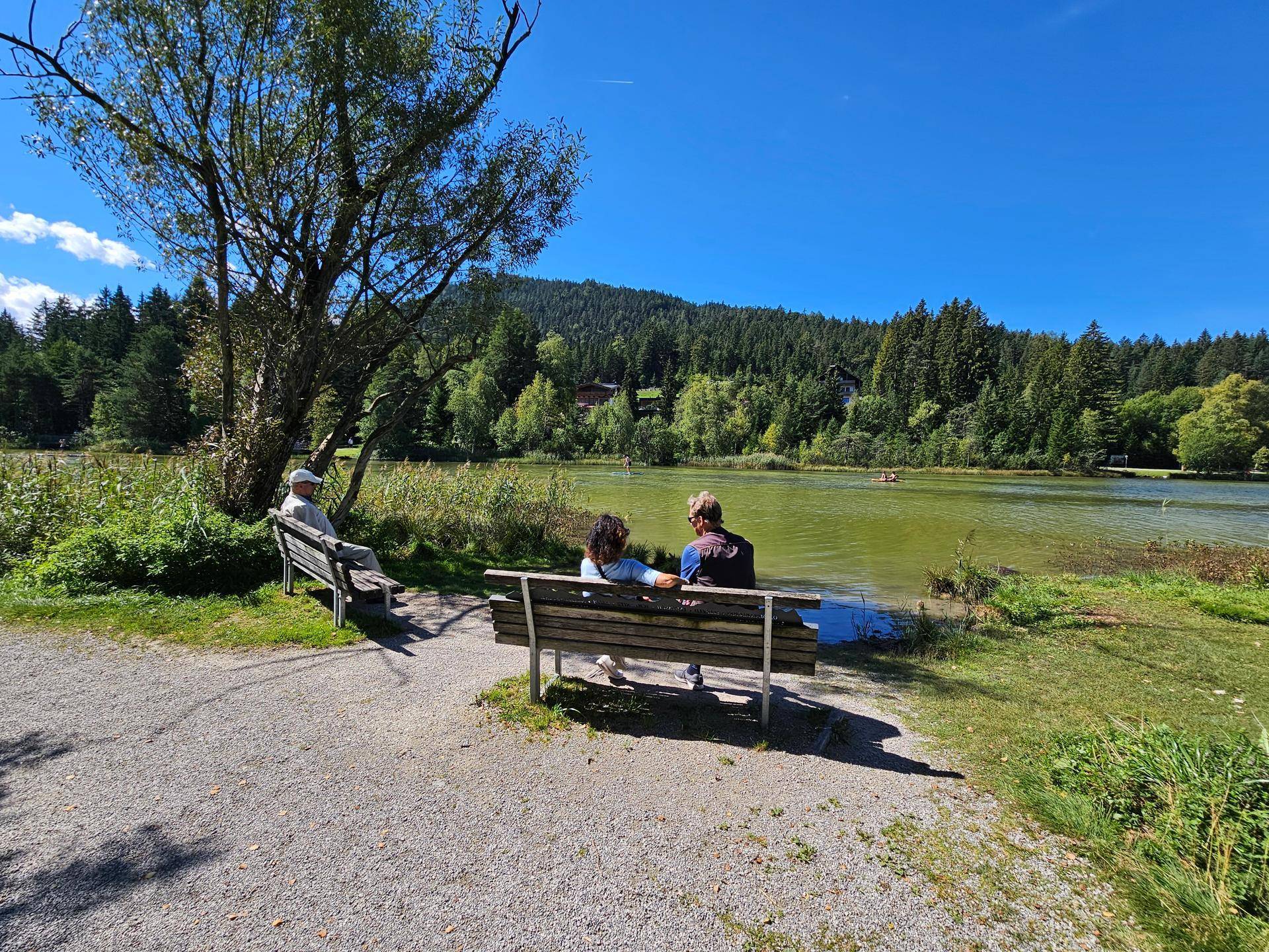 Menschen sitzen auf einer Bank am Ufer eines Sees mit bewaldeten Bergen im Hintergrund an einem sonnigen Tag.