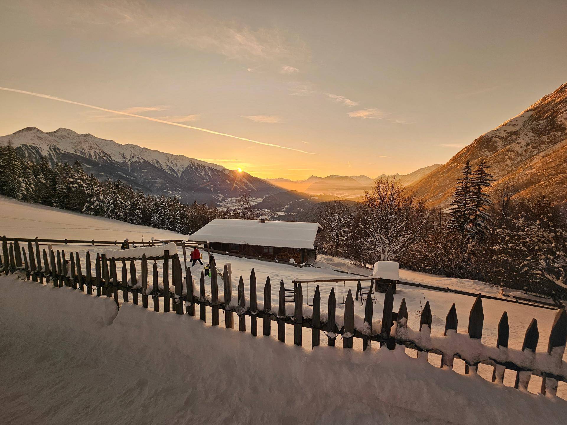 Atemberaubender Sonnenuntergang über einem verschneiten Tal mit einer Holzhütte und Bergen.