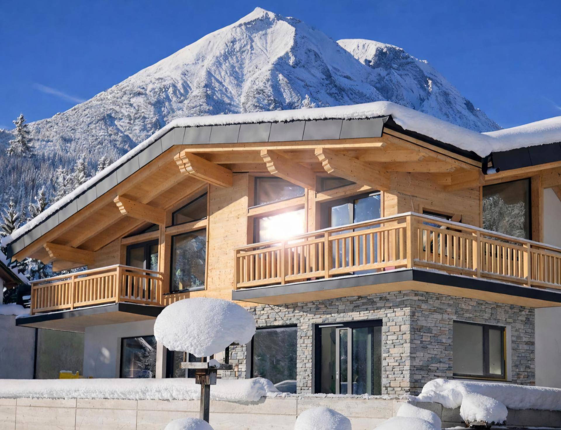 Modernes Holzhaus mit Balkonen und Steinverkleidung vor einer beeindruckenden Bergkulisse im Winter.