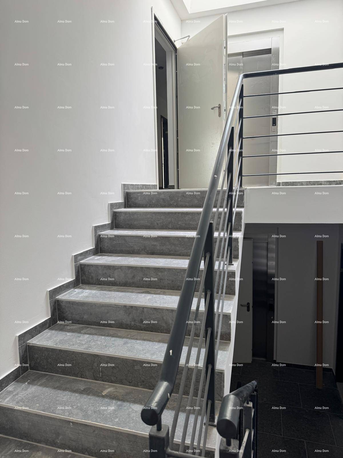 Treppenhaus mit modernen grauen Stufen und schwarzem Geländer, neben einem Aufzug.