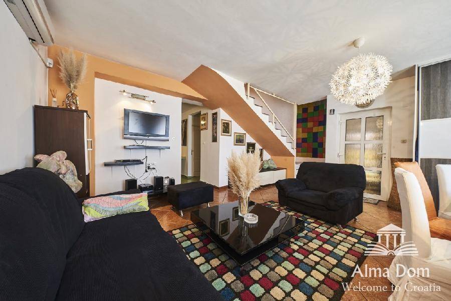 Heller Wohn- und Essbereich mit Treppe, Sofa und Fernseher an der Wand auf Fliesenboden.