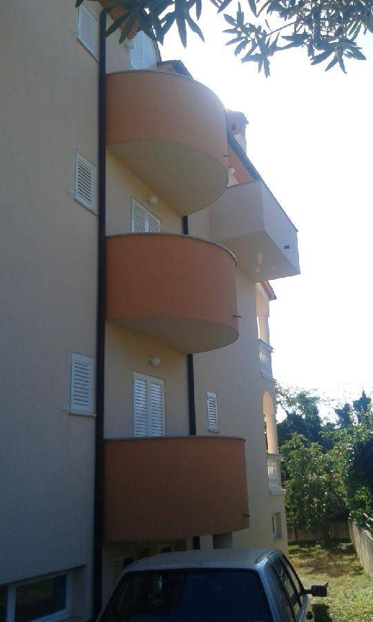 Seitenansicht eines Gebäudes mit mehreren geschwungenen Balkonen und weißen Fensterläden unter hellem Sonnenlicht.
