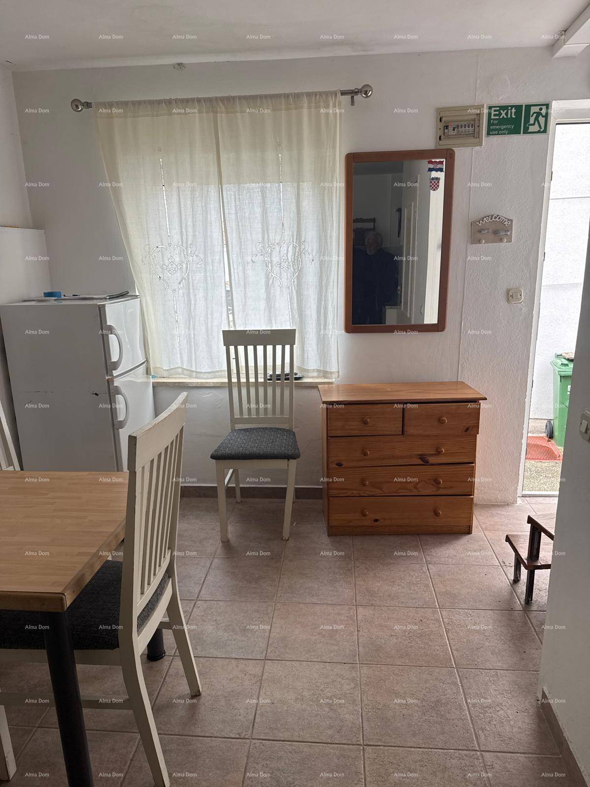 Heller Raum mit Fliesenboden, einem Kühlschrank, Kommode und einem Fenster mit Vorhängen.