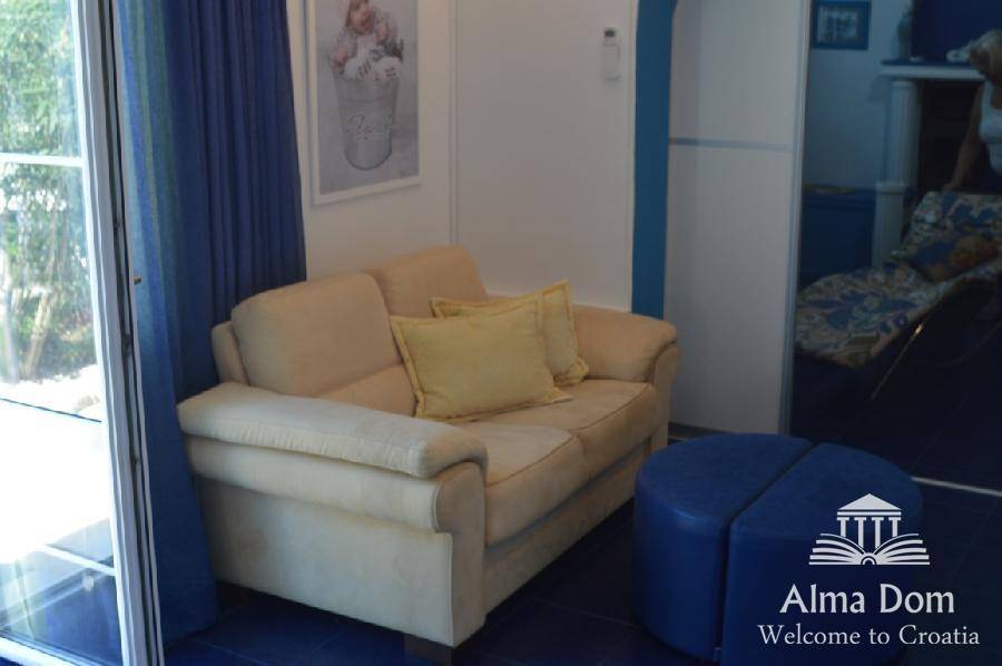 Gemütliches Wohnzimmer mit beigem Sofa und blauem Hocker, mit Zugang nach draußen.
