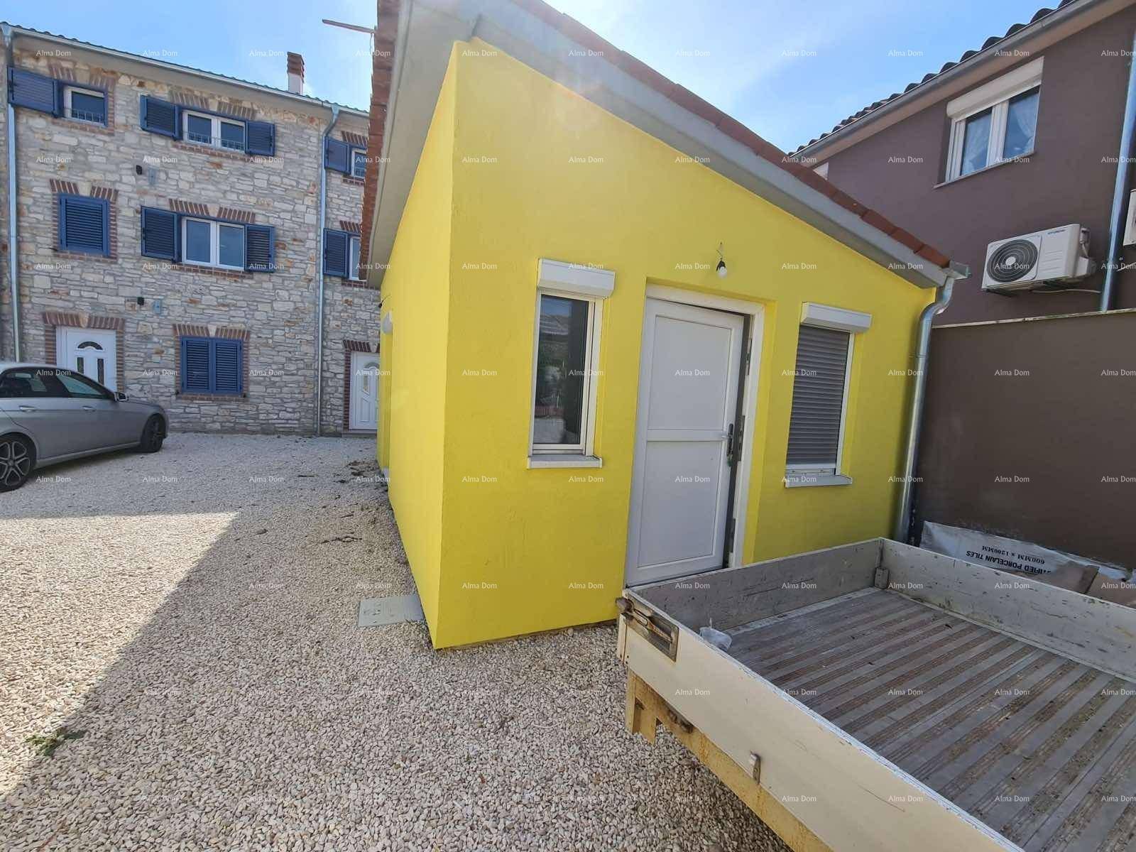Gelbes Gebäude mit weißer Tür und Fenster, umgeben von Kies, neben einem traditionellen Steinhaus.
