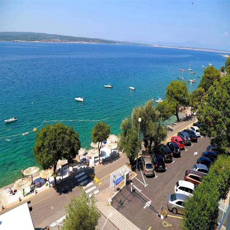 Blick von oben auf einen Parkplatz am Meer, mit Strandabschnitt und zahlreichen Booten im Wasser.