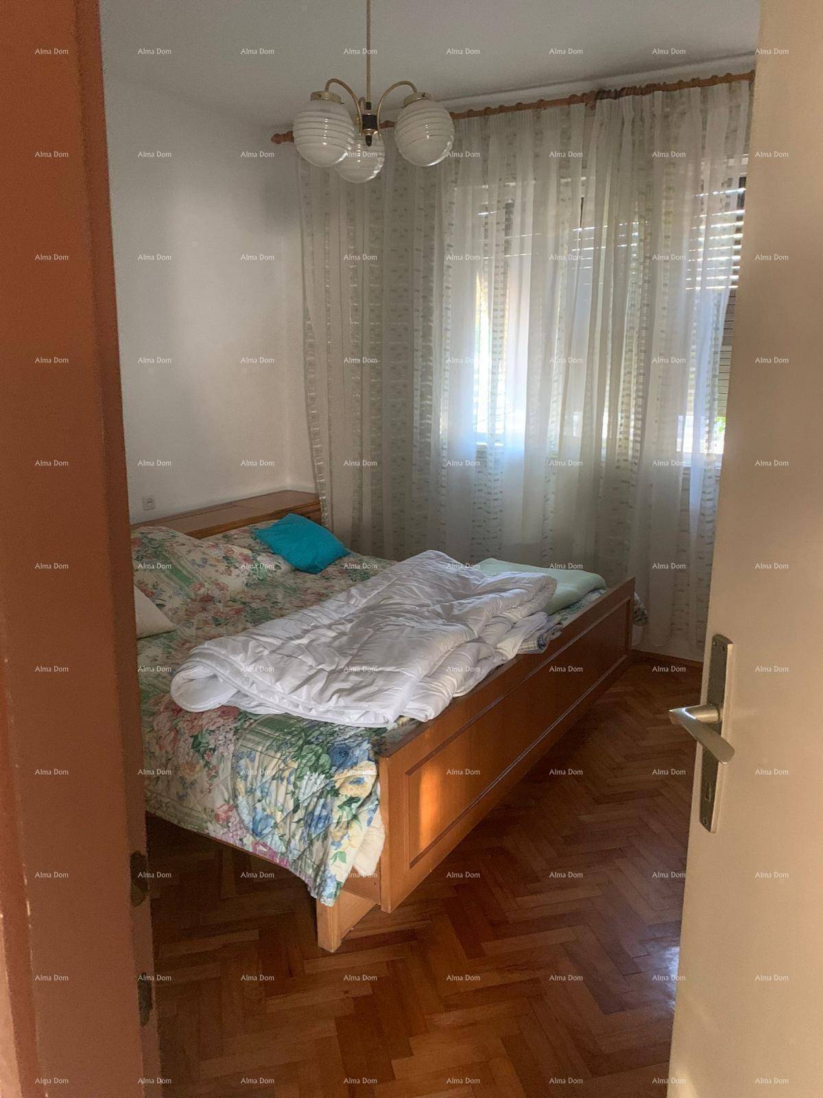 Schlafzimmer mit einem Doppelbett, hellem Parkettboden und einem Fenster mit transparenten Vorhängen.
