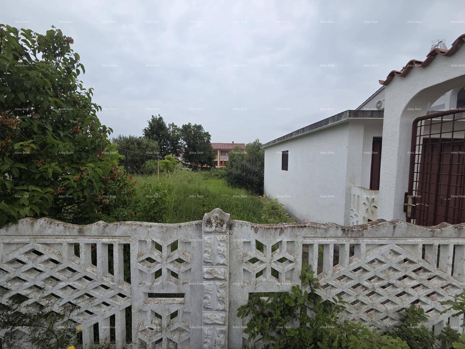Blick über einen dekorativen Betonzaun auf ein verwildertes Grundstück mit einem weißen Gebäude im Hintergrund.
