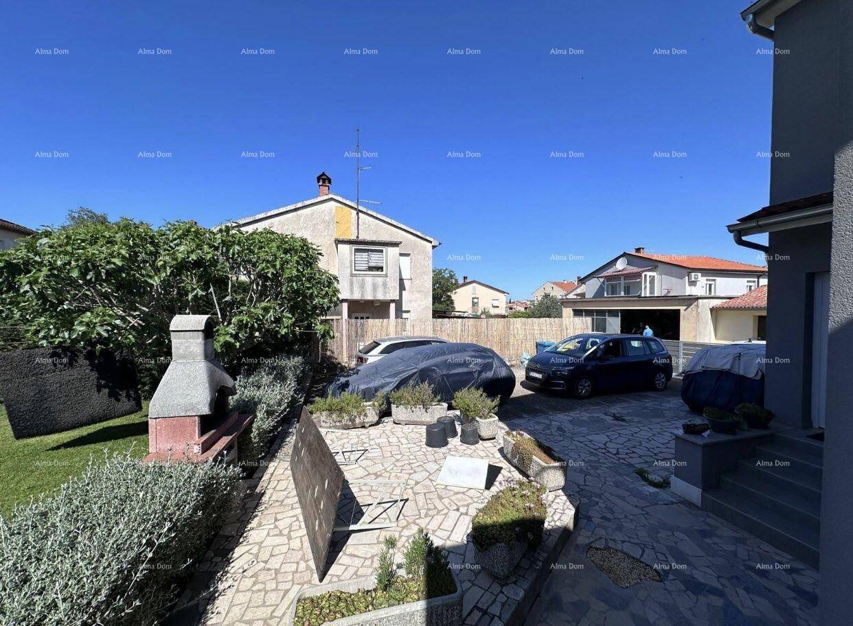 Garten und gepflasterter Bereich mit Grill, Pflanzenkübeln und parkenden Autos.