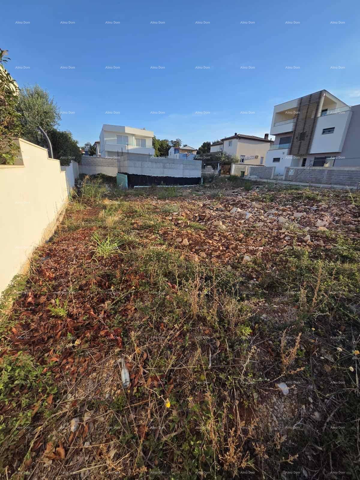 Unbebautes Grundstück mit trockener Vegetation und einer weißen Mauer, umgeben von Neubauten.