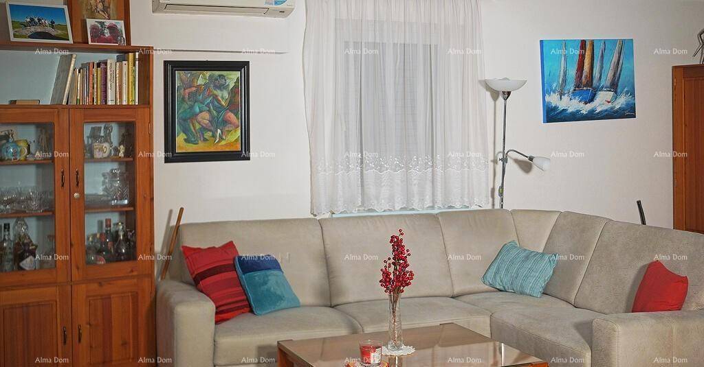 Gemütliches Wohnzimmer mit beigem Ecksofa, Bücherregal und Klimaanlage, hell durch ein Fenster.