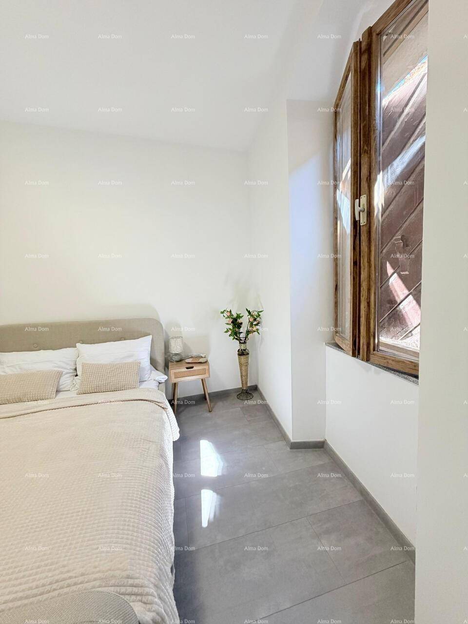 Helles Schlafzimmer mit Doppelbett, Holzbalkenfenster und modernem Fliesenboden, schlicht eingerichtet.