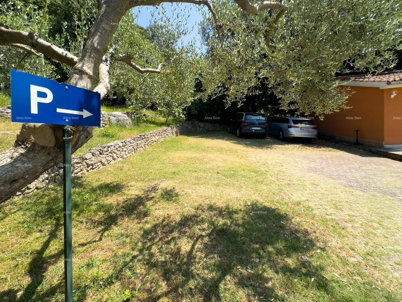 Parkplatzbereich mit Kies und Gras, umgeben von Bäumen und einer Steinmauer, mit einem Parkschild.