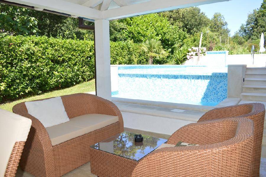 Gemütliche überdachte Terrasse mit Rattanmöbeln und Blick auf den Poolbereich und die grüne Umgebung.