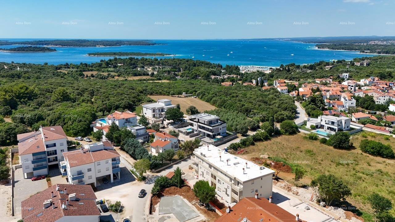 Luftaufnahme einer Küstensiedlung mit Wohngebäuden, viel Grün und dem Meer im Hintergrund unter blauem Himmel.
