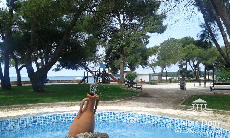 Grüner Park mit Springbrunnen, Spielplatz und Blick auf das Meer im Hintergrund, ideal für Familien.