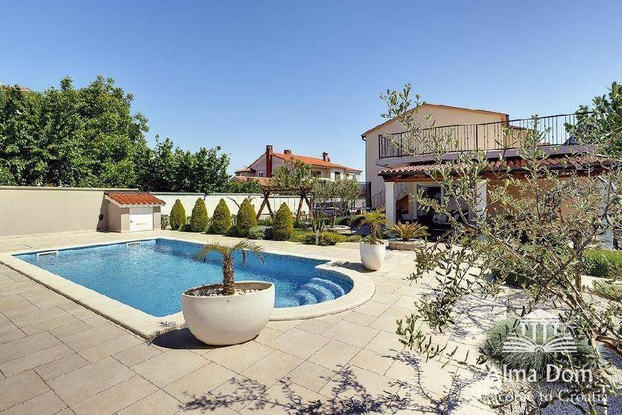 Sonniger Poolbereich mit gepflasterter Terrasse, mediterranen Pflanzen und Blick auf das Haus.