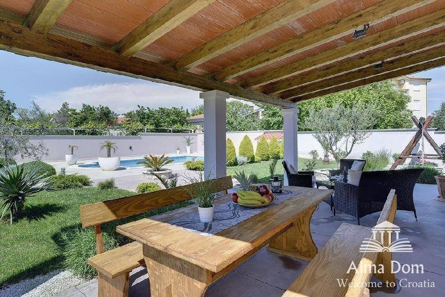 Geräumiger Essbereich im Freien unter einer Holzpergola mit Blick auf den Pool und Garten.