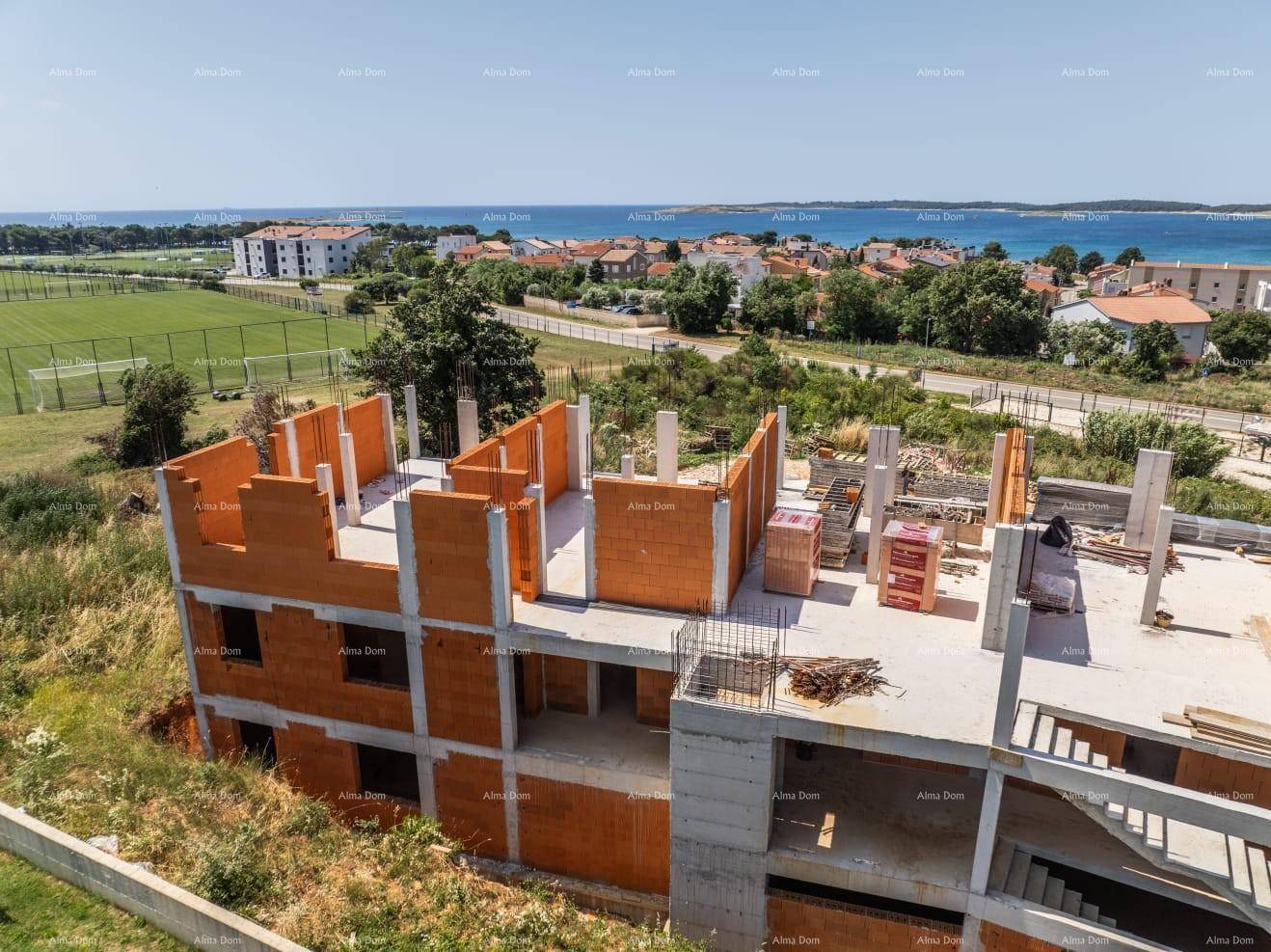 Luftaufnahme der Baustelle aus niedrigerer Höhe mit Sportfeldern und Meerblick im Hintergrund.