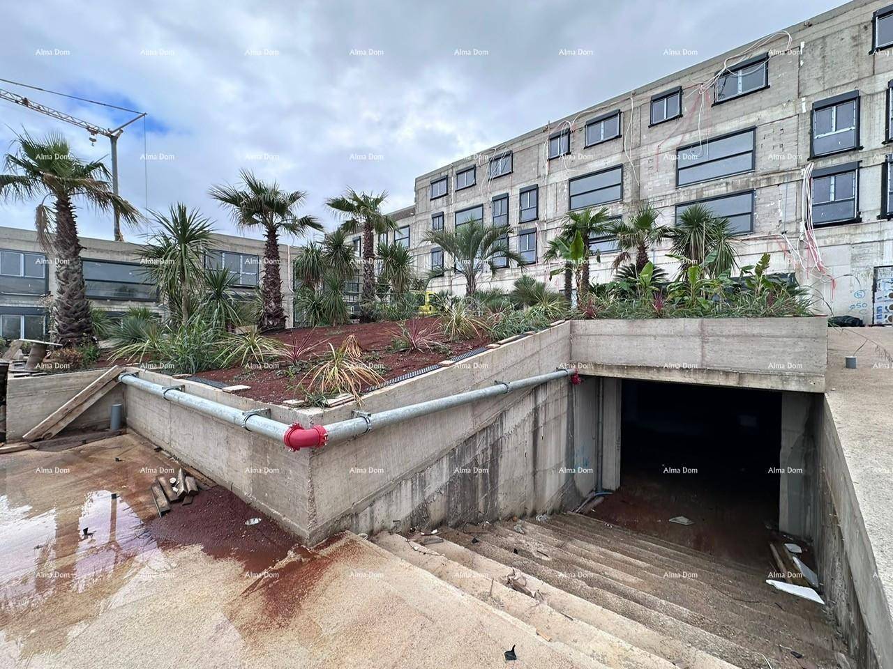 Außenansicht eines im Bau befindlichen Gebäudes mit Tiefgaragenzufahrt und Palmen.