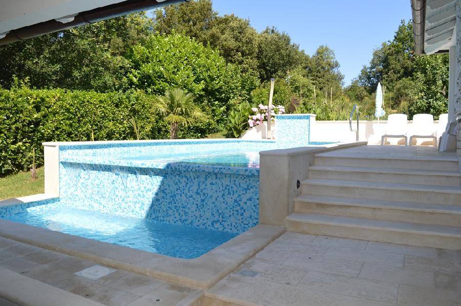 Moderner Pool mit Wasserfallfunktion und angrenzender Sonnenterrasse, umgeben von üppigem Grün.