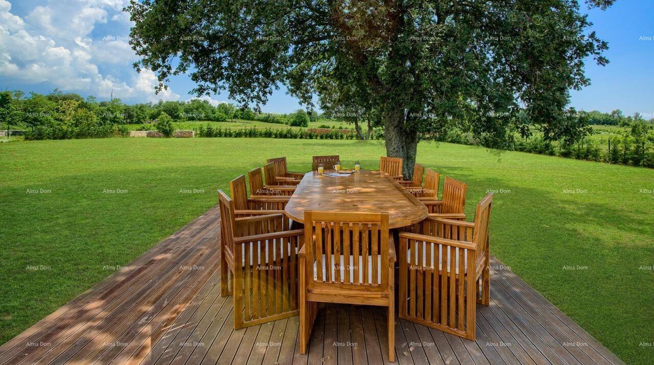 Großer Holztisch mit Stühlen auf einer Holzterrasse unter einem Baum, umgeben von grünen Feldern.