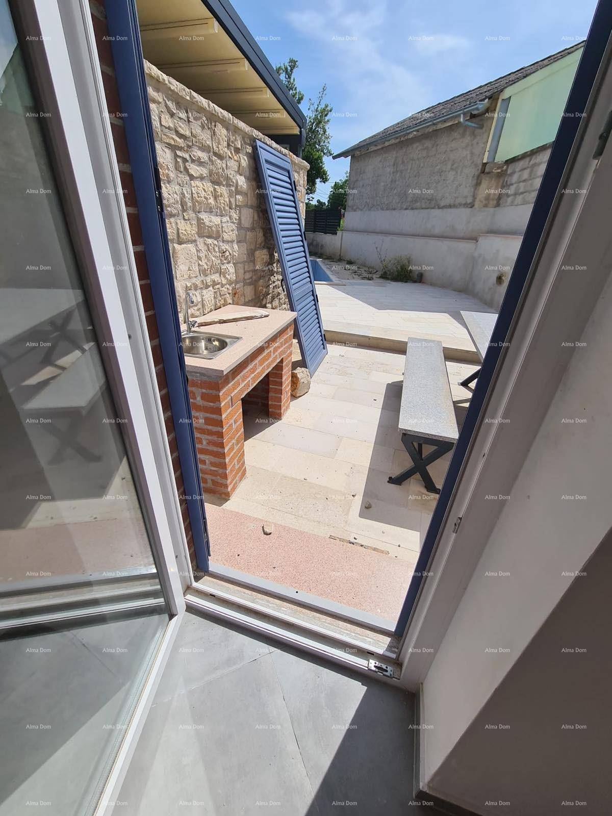 Blick vom Innenraum auf eine gepflasterte Terrasse mit gemauertem Grillbereich.