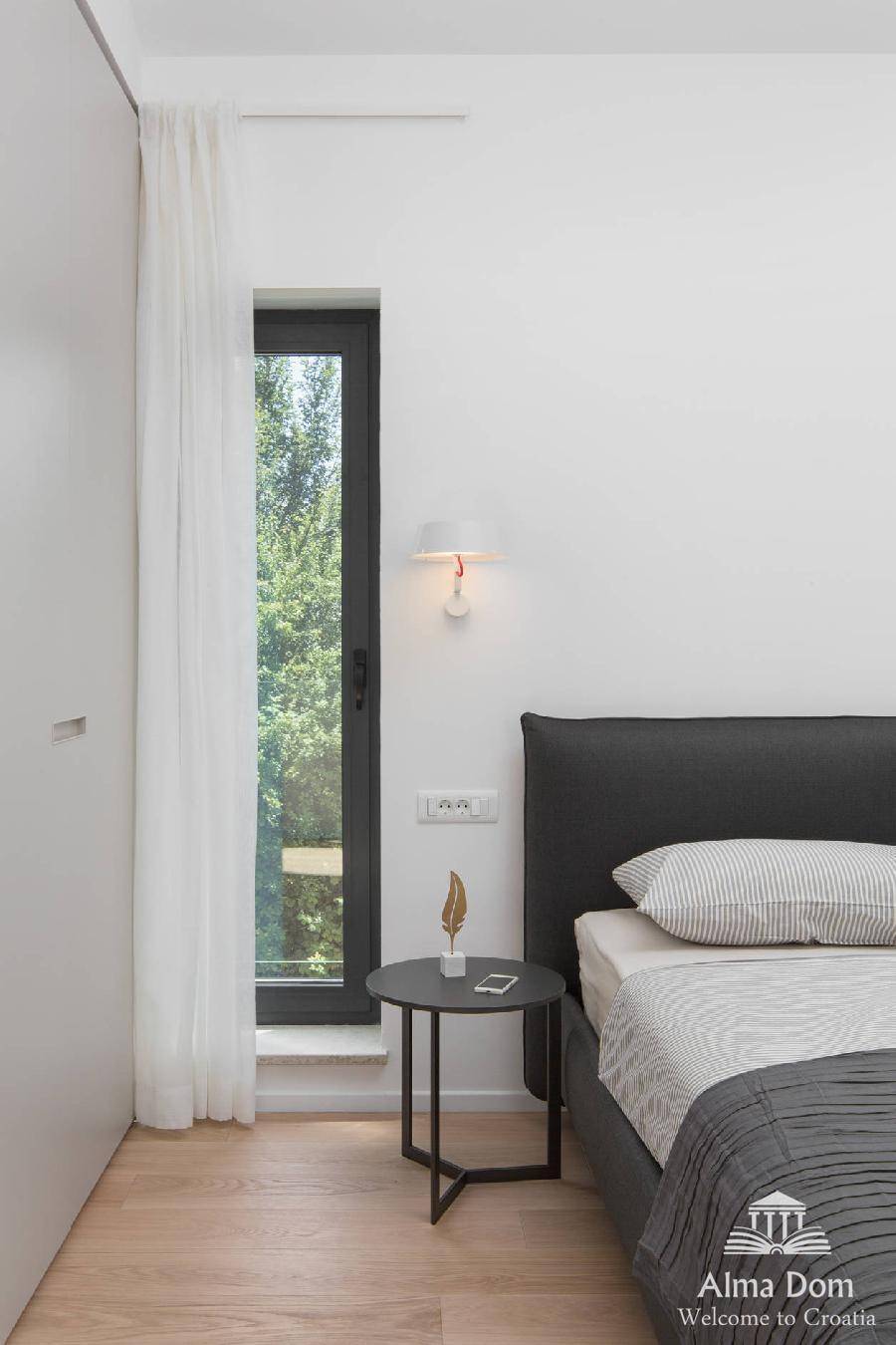 Helles Schlafzimmer mit Blick ins Grüne durch ein hohes Fenster und minimalistischer Einrichtung.