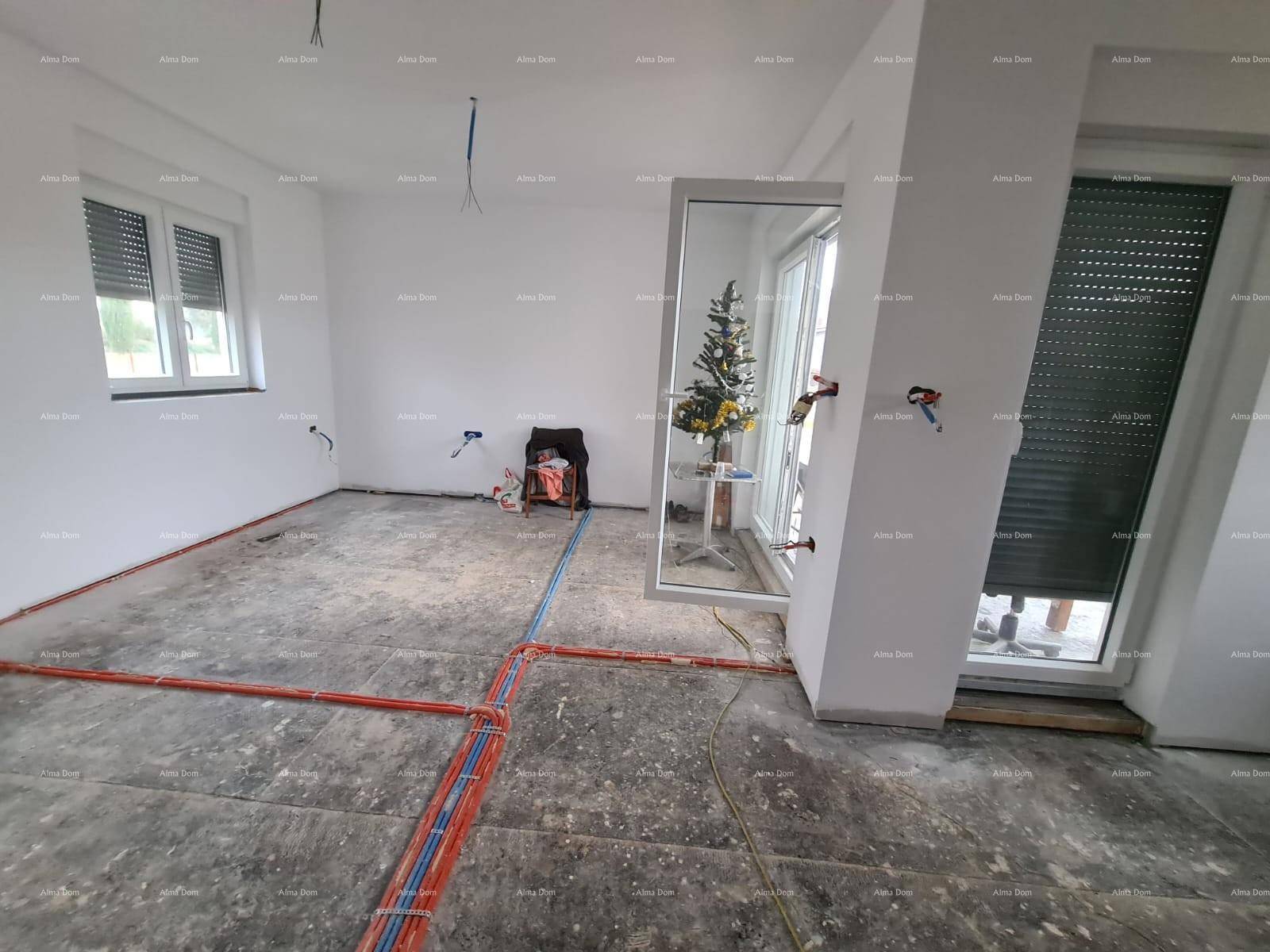 Unfertiger Innenraum mit sichtbaren Fußbodenheizungsrohren, Fenster mit Rollladen und viel Tageslicht.