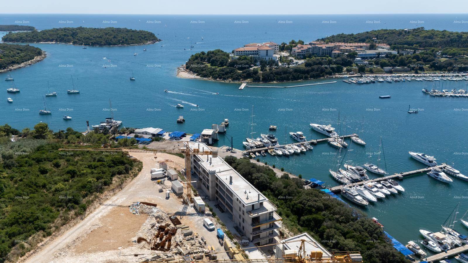 Luftaufnahme einer Baustelle mit einem im Bau befindlichen Gebäude und einem Yachthafen mit vielen Booten.